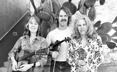 Three people stand in front of a cactus. From left to right, a person holds a guitar, another with a violin, and a third holds a mandolin. All are casually dressed and looking at the camera with neutral expressions. Black and white image.