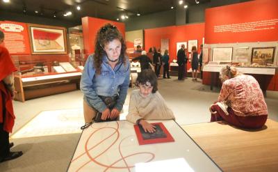 A woman and a child interact with a display at a museum exhibit. The child is touching a screen with red lines. Other visitors are in the background, and various artifacts and information panels are on the walls.