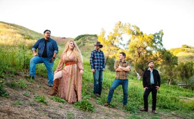 Five people stand on a grassy hillside. The group includes a person in a long dress and boots, and others in casual attire with jeans. One wears a cowboy hat. Trees and rolling hills are in the background under a clear sky.