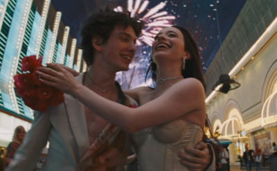 A young couple embracing and smiling joyfully under a lit-up sky filled with fireworks. They are wearing formal attire, with the girl holding a bouquet of red roses. The backdrop includes neon lights and an arcade-like setting, suggesting celebration.