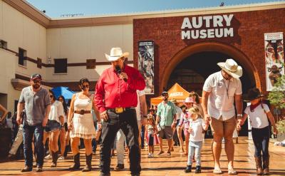 A group of people in cowboy hats and boots are line dancing outside the Autry Museum on a sunny day. The scene is lively, with a mix of adults and children participating. The museum's brick facade is visible in the background.
