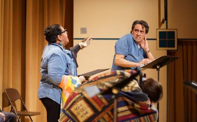 A dramatic stage scene with two actors. One gestures emotionally towards a man behind a lectern, who appears surprised. A colorful patchwork quilt drapes over another person seated nearby. Both actors wear casual blue shirts.
