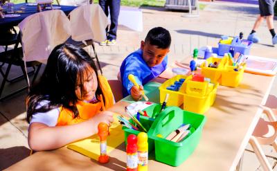 Two children sitting at a table outdoors, wearing aprons and painting. The table is filled with art supplies, including brushes and jars of paint in various colors. The background shows more tables and people.