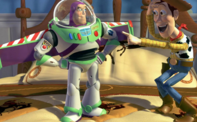 A cowboy toy points excitedly at an astronaut action figure standing on a bed. The bed has a cowboy-themed blanket. The scene takes place in a child's room.