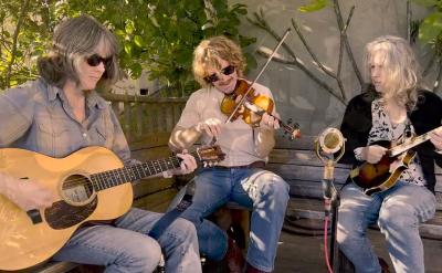 Three musicians sit on a wooden bench outdoors, surrounded by greenery. One plays an acoustic guitar, another a violin, and the third a mandolin, with a microphone set up in front of them.