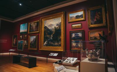 A gallery with framed paintings on red walls displays various landscapes and portraits. Glass cases exhibit silver artifacts. Wooden benches provide seating. The room is softly lit, enhancing the art's details.