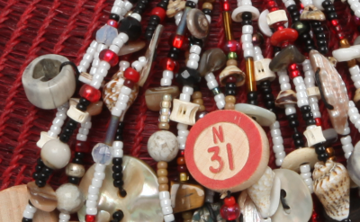 A close-up of numerous colorful beads on a basket with various charms, including circular pieces with numbers "N 31" and "O 75." The necklaces rest on a textured red surface.