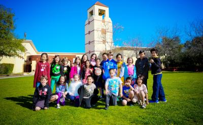 A group of children pose and smile together on a lawn in front of a school building with a tower under a bright blue sky.