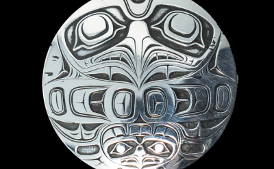 A round silver medallion featuring intricate, symmetrical indigenous art with bold lines and stylized animal or mythical face designs engraved on the surface. The background is black.