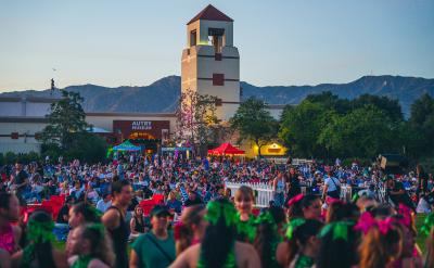 A large crowd gathers outdoors in front of the Autry Museum at dusk, with mountains in the background and a tower visible. People sit on chairs and blankets; festive decorations and tents add to the lively atmosphere.