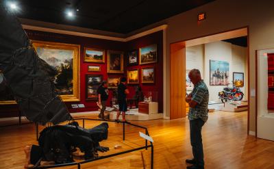 A man stands in a museum gallery with red walls, viewing various paintings and exhibits. Two people are observing artworks on the left. A motorcycle is displayed in a lit adjoining room.