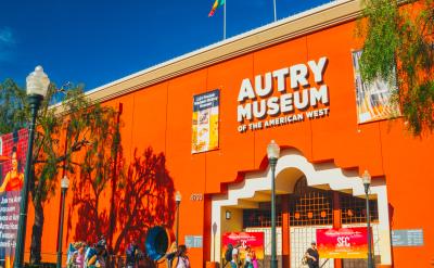Visitors walk outside the bright orange facade of the Autry Museum of the American West on a sunny day. The building features banners, three flags on top, and large white signage above the entrance.