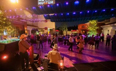 People in western attire dance under string lights at an outdoor evening event at the Autry Museum. Musicians perform on stage in the foreground; trees and buildings are decorated with festive lights.