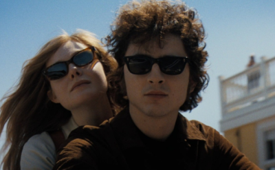 A young woman and man wearing dark sunglasses ride together on a motorcycle under a clear blue sky, with a building in the background. The woman leans on the man's shoulder, both looking calm and composed.
