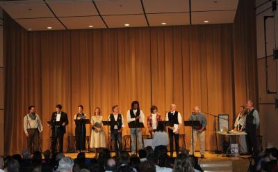 A group of ten performers stands on a stage in front of an audience, facing forward. Most are holding scripts. The stage background is tan curtains, and a person stands to the left of a table with audio equipment on the right side.