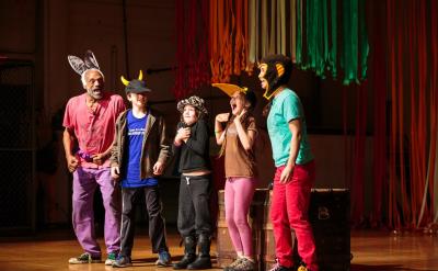 Five people, including children and adults, stand on stage wearing colorful animal hats and costumes. They are smiling and laughing, with colorful streamers hanging in the background.