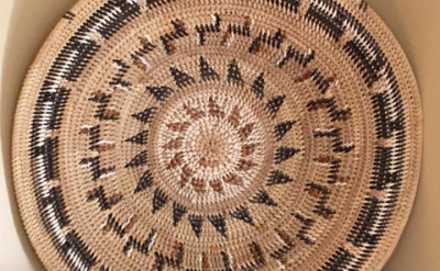 A large, round woven basket with intricate geometric patterns in natural, black, and brown colors is displayed upright against a beige wall on top of a dark wooden surface.