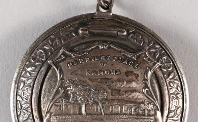 A round silver medallion with an engraved image of a house and trees, inscribed with "Marriage Place of Ramona" and "San Diego, Cal," with decorative patterns around the border and a loop at the top for attachment.
