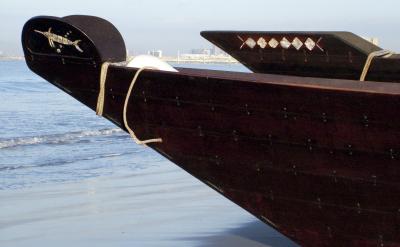 Close-up of the bow of a traditional wooden dhow boat on a beach, with rope detail and geometric decorations; city buildings are visible in the distant background across the water.