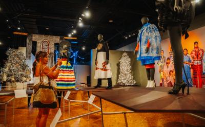 A woman takes a photo of mannequins dressed in colorful, elaborate outfits on display in a museum exhibit with dramatic lighting and various artwork in the background.