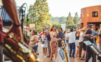 People dance outdoors in a lively, sunlit plaza, surrounded by trees and buildings. Musicians with brass instruments play in the foreground, and many couples enjoy dancing together in the background.