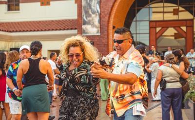 A joyful crowd dances outdoors in front of a building. A smiling woman with curly hair and a man in a colorful shirt are dancing together in the foreground, surrounded by other people enjoying the lively atmosphere.