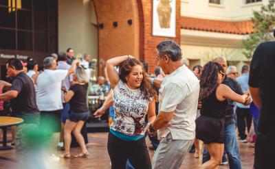 People dancing joyfully in an outdoor plaza, with a band playing in the background. The scene is lively, with men and women smiling and enjoying the music on a sunny day.