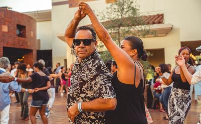 A man and woman dance together in an outdoor courtyard, surrounded by other people who are also dancing. The man wears sunglasses and a floral shirt, while the woman smiles as she spins under his arm.