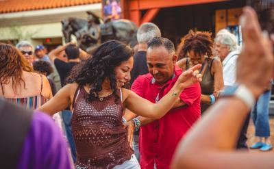 A woman and man dance joyfully outdoors in a crowd, surrounded by people smiling and enjoying the lively atmosphere. The woman raises her arm while the man smiles, closely following her moves.