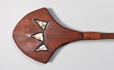 A wooden paddle with a wide, flat blade featuring three triangular mother-of-pearl inlays, and a long, smooth handle wrapped with woven material near the blade.