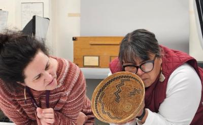 Two women examine a woven basket with intricate patterns; one woman closely inspects the basket while the other leans in with interest. They appear to be in a workspace with papers and objects nearby.