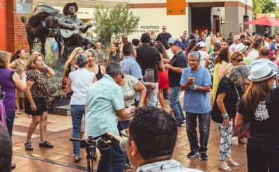 A large group of people dances and socializes outdoors in a plaza, with a musician statue playing guitar in the background and trees and buildings surrounding the lively crowd.