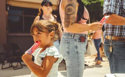 A young girl with brown hair eats a red popsicle while standing outside in a line with adults on a sunny day. She looks off to the side, and people around her are casually dressed in summer clothing.