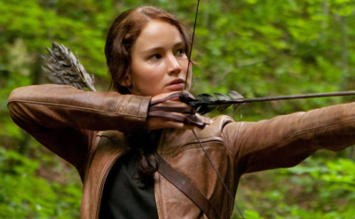 A young woman with brown hair, wearing a brown jacket and quiver, draws a bow and aims an arrow in a lush green forest.