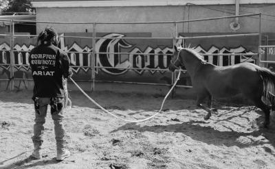 A person wearing chaps and a "Compton Cowboys" shirt holds a lunge line, training a horse in a sandy round pen with a large mural on the wall behind them.