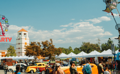 People enjoy a lively outdoor block party with vintage cars, a Ferris wheel, tents, and trees. The Autry Museum logo and event name appear on the left against a blue, partly cloudy sky.
