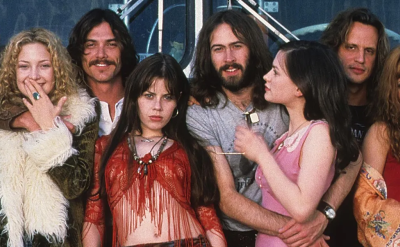 A group of people in 1970s-style clothing pose together, some smiling and some serious, in front of a tour bus. They appear to be members of a rock band and their entourage.