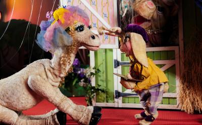 A costumed character resembling a lamb wearing a colorful hat with flowers and veil kneels on a red carpet, facing a smaller puppet in yellow clothes and bunny ears, set in a whimsical indoor scene.