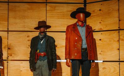Four mannequins wear classic Wild West costumes, including hats, bowties, vests, long coats, skirts, holsters, and bandanas, standing in front of a wooden panel wall.