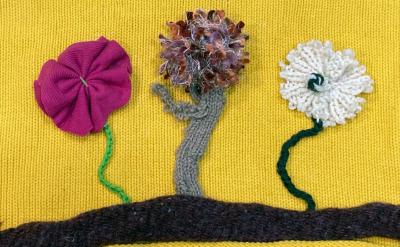 Three textured flowers made from fabric and yarn are attached to a yellow background. The flowers are pink, brown, and white, each with a green stem, standing on a brown horizontal line.