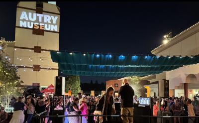 A large crowd attends an outdoor nighttime event at the Autry Museum, with string lights overhead, people mingling, and a DJ booth in the foreground. The museum’s name is projected on a building wall.