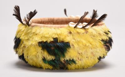 A round woven basket decorated with yellow and green feathers, brown feathers sticking up at the rim, and white shell-like ornaments; the background is plain light gray.