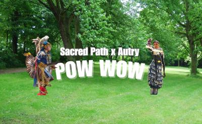 Two Native American dancers in traditional regalia stand on green grass in a park with trees. Bold white text reads "Sacred Path x Autry POW WOW" across the center of the image.