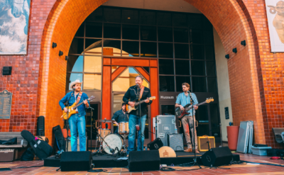 A band performs live music on an outdoor stage beneath the large arched entrance of the Autry Museum, with instruments, speakers, and museum signage visible in the background.