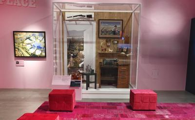 A museum exhibit in a pink room displays a glass case with a typewriter, a table, a lamp, framed photos, and landscape images. Red cushioned seats and a pink-patterned rug are in front of the display.