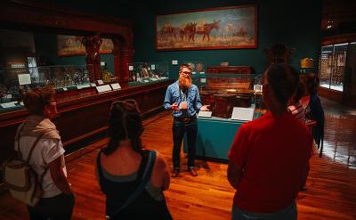A museum guide speaks to a small group of visitors in a dimly lit exhibit room with wooden floors, display cases, and a large painting of horses on the wall.