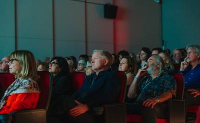 A diverse group of people sits in red theater seats, attentively watching something off-camera. The audience appears focused and engaged, with dim lighting highlighting their faces.