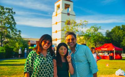 Three people stand smiling on a sunny lawn with a white tower and trees in the background. The girl in the middle makes a peace sign. Red tents and blue sky are visible in the distance, giving a festive atmosphere.