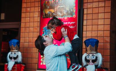 A woman lifts a smiling young girl in a red sweater and glasses, touching noses, in front of a festive display with toy soldier figures and bright red decorations.