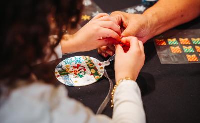 A child and an adult use their hands to decorate a round ornament with colorful stickers and designs on a dark table, suggesting a festive craft activity.